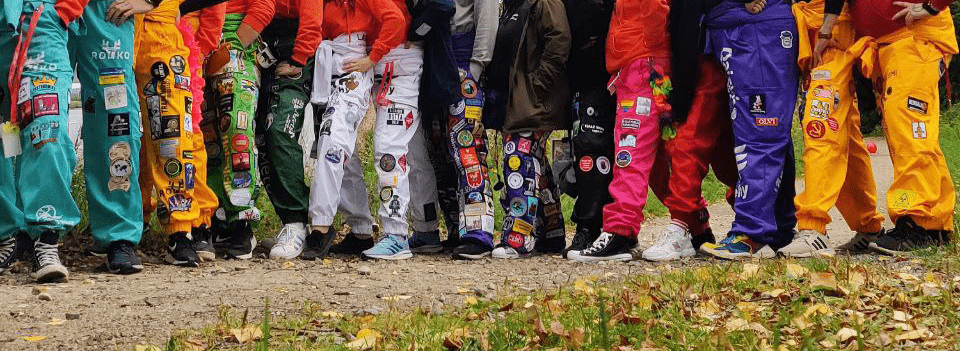 kuva, jossa eriväriset haalarit päällä olevat ihmiset seisovat rivissä. Kuva rajattu näyttämään vain vyötäröstä alaspäin, eli pelkät haalarijalat näkyvät.