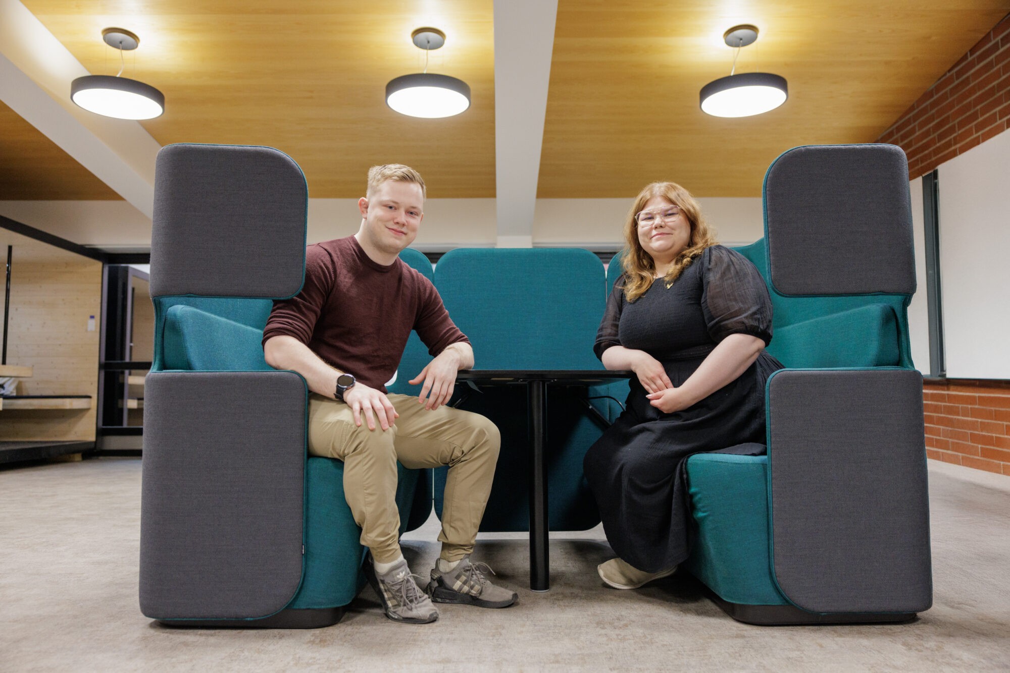 Kaksi henkilöä istuu vastakkain modernissa työtilassa, pöydän ääressä sinivihreissä sohvaryhmissä.